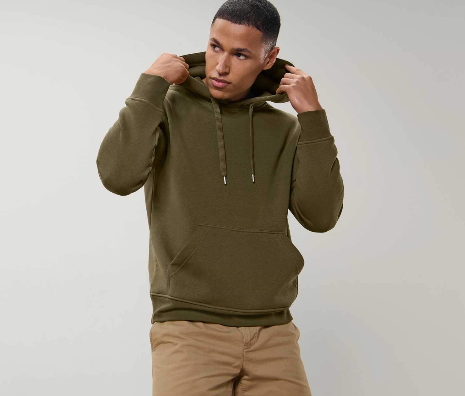 Muž si upravuje kapucňu mikiny s kapucňou olivovej farby. Má na sebe béžové šortky.