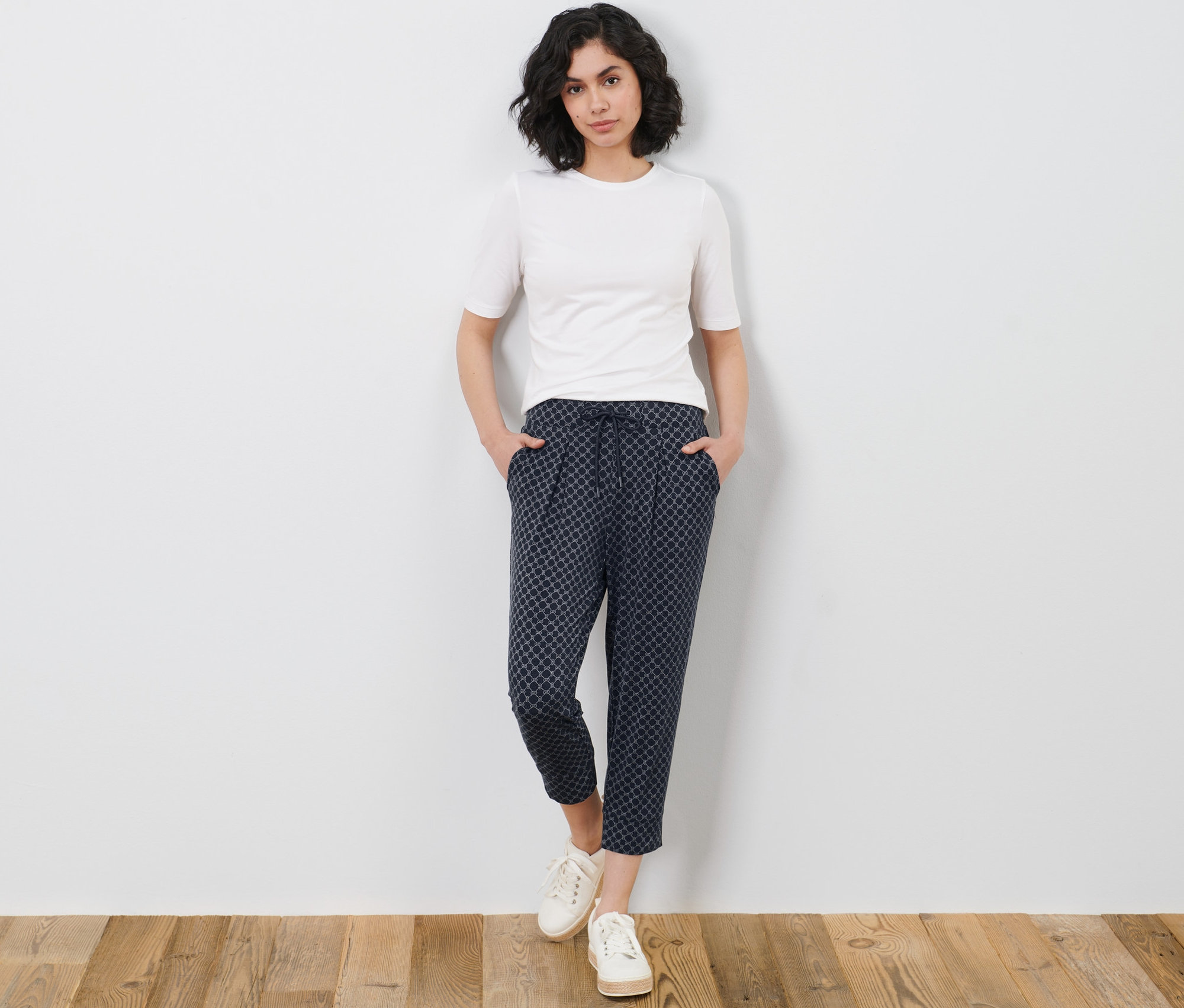 Žena pózuje v bielom tričku a trojštvrťových džersejových nohaviciach s potlačou.