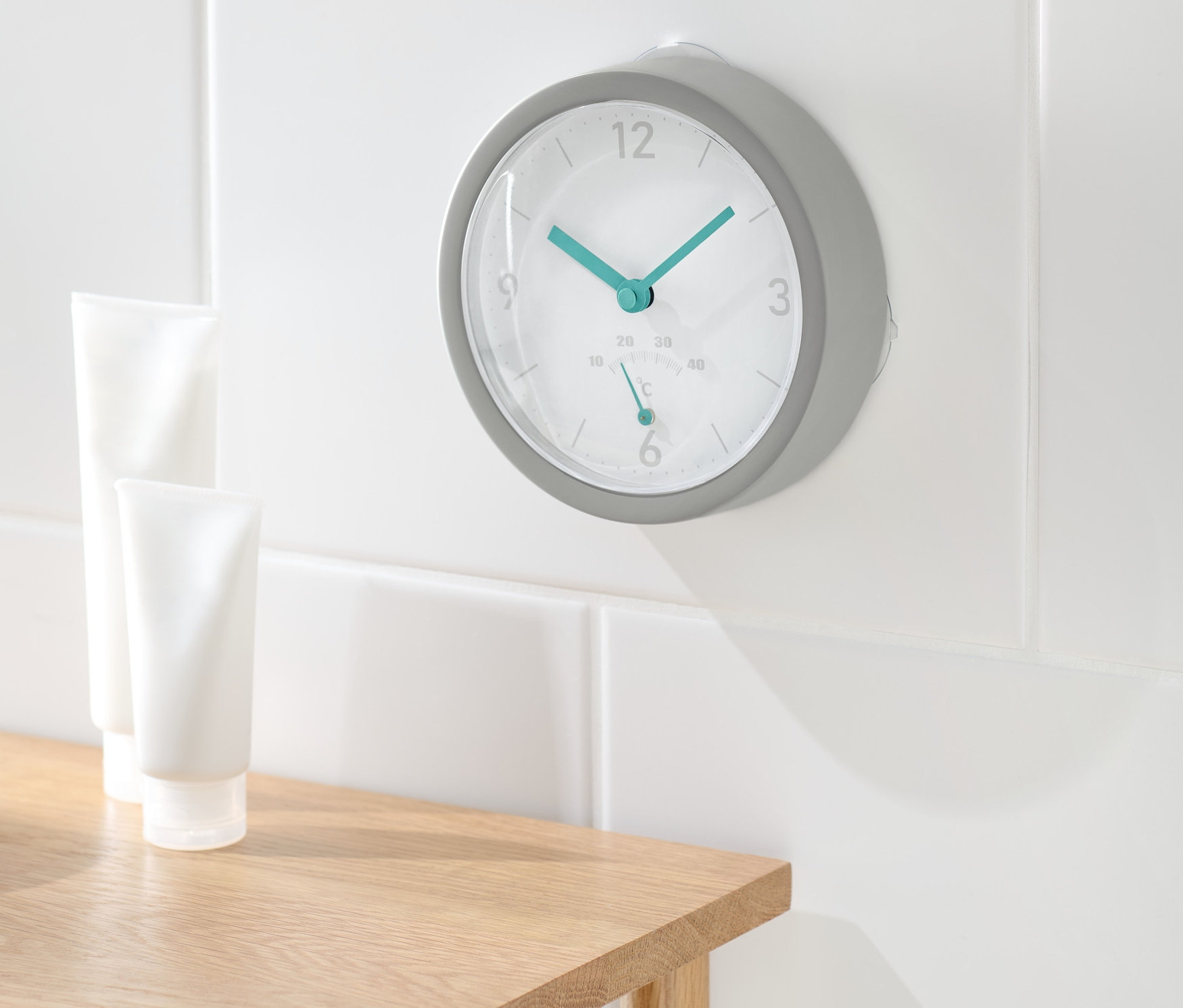 Na bielej stene kúpeľne visia bezdrôtové nástenné hodiny s teplomerom. Na drevenom stolíku stoja dve biele tuby.