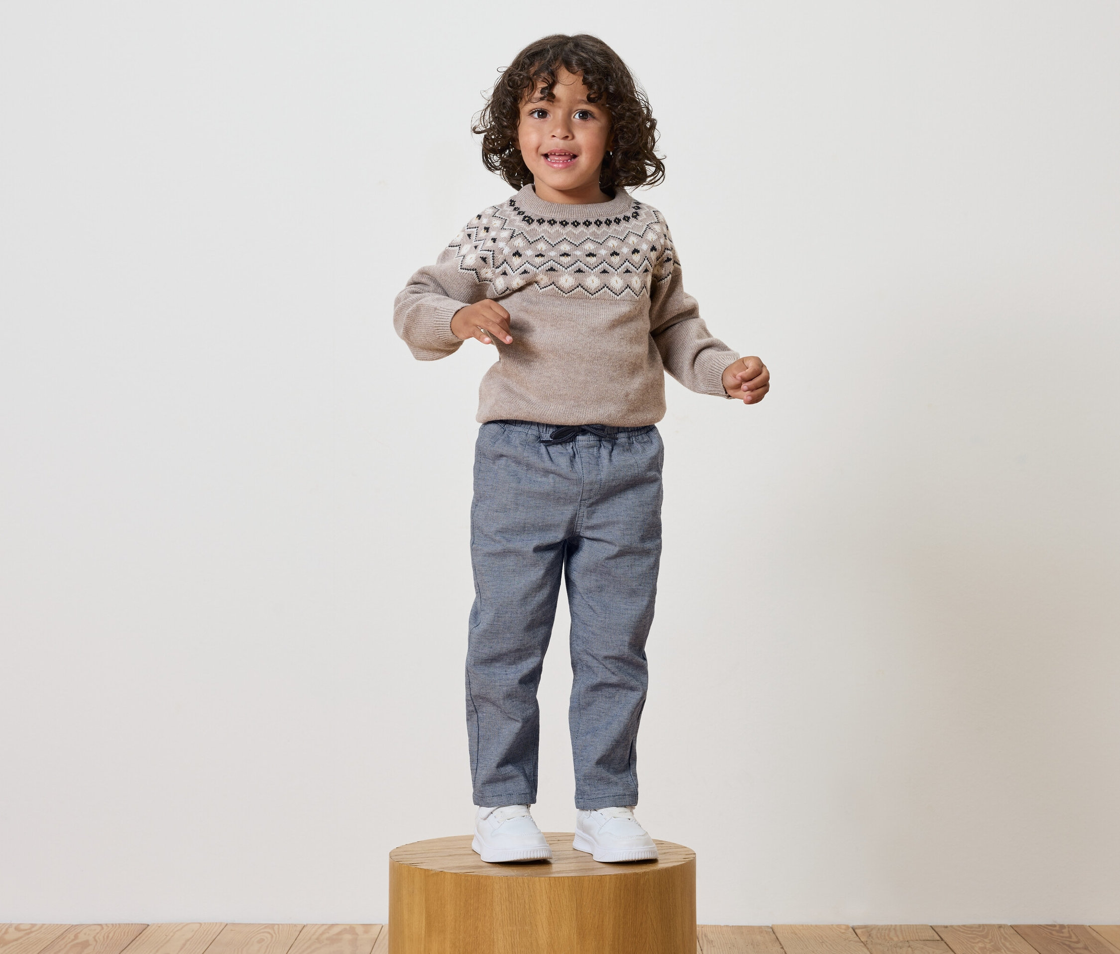 Chlapec s kučeravými vlasmi stojí na drevenom podstavci, má na sebe sveter so vzorom a modré nohavice.