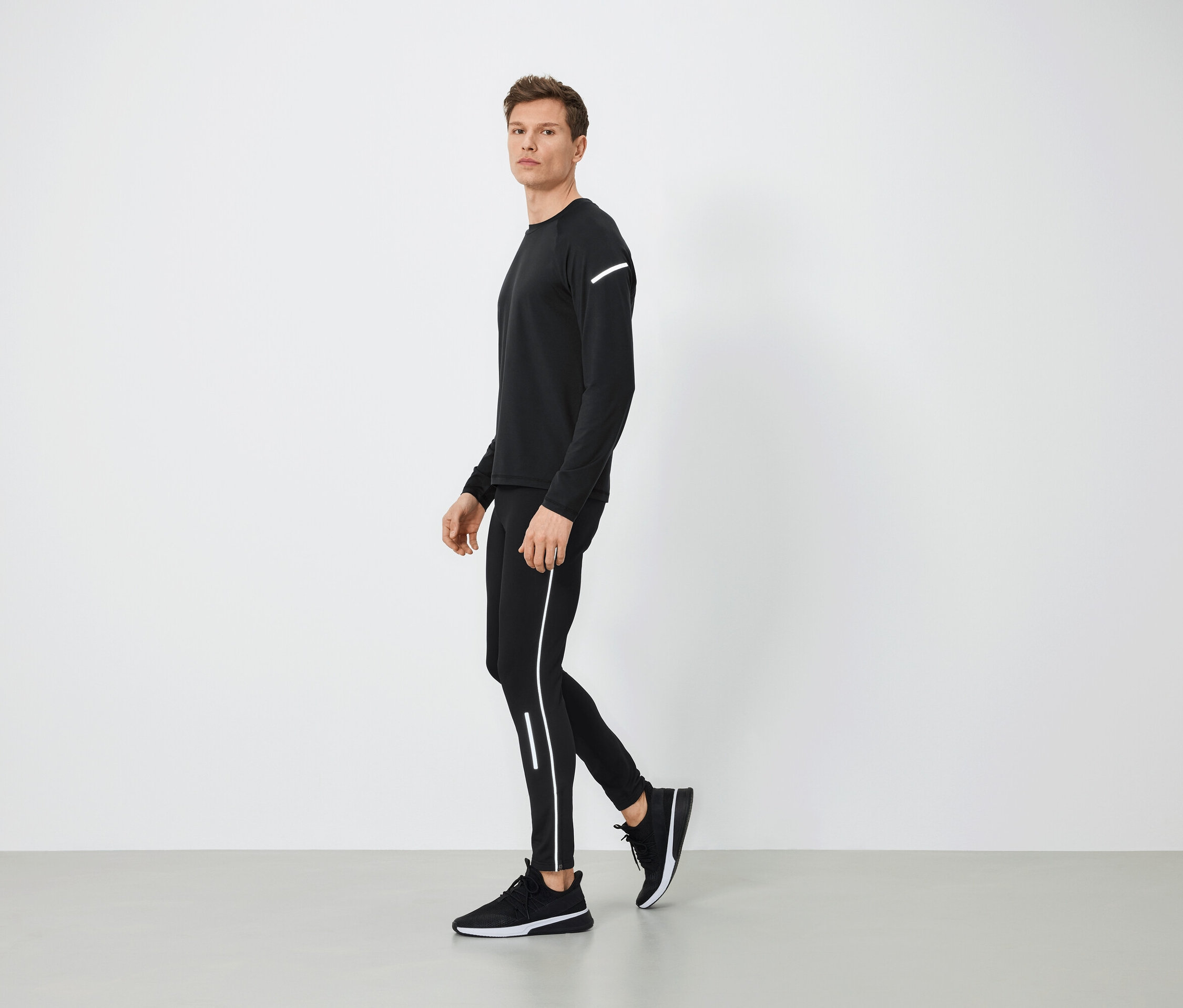 Muž v čiernom športovom oblečení s reflexnými prvkami kráča po šedej podlahe.