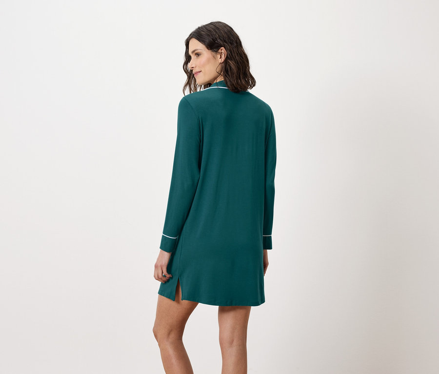 Žena stojí chrbtom a má na sebe smaragdovo zelenú nočnú košeľu s dlhými rukávmi a bielym lemovaním.