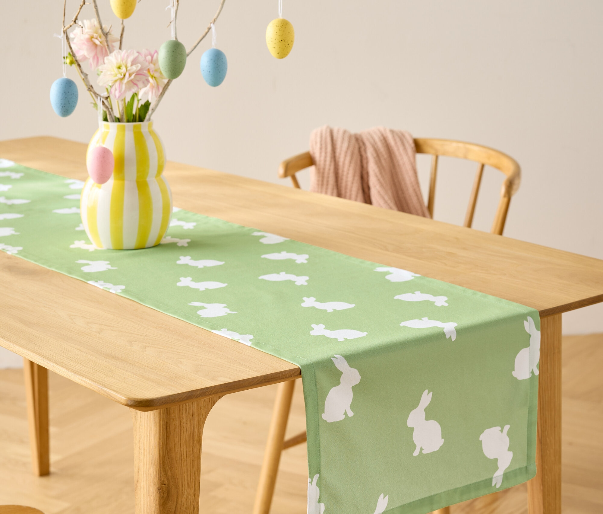 Jedálenský stôl so svetlozeleným behúňom s bielymi motívmi zajačikov, váza s kvetmi a dekorácia s veľkonočnými vajíčkami.