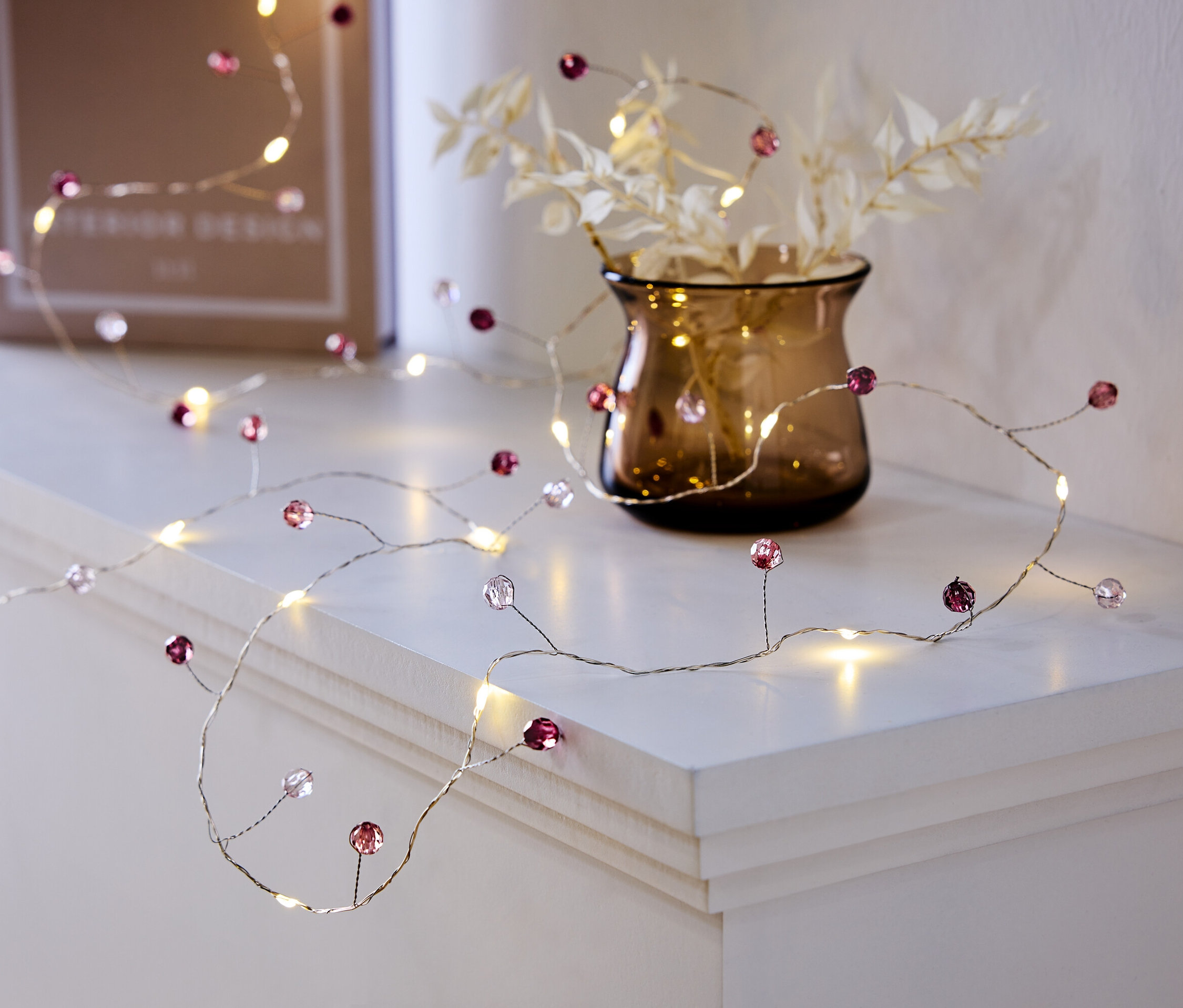 Dekoratívna svetelná reťaz s fialovými a priehľadnými korálkami leží na bielej polici vedľa vázy s rastlinami a knihy.