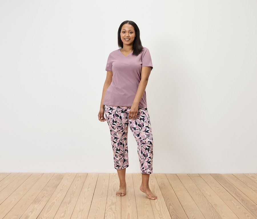 Žena stojí a má na sebe fialové tričko a kvetinové nohavice.