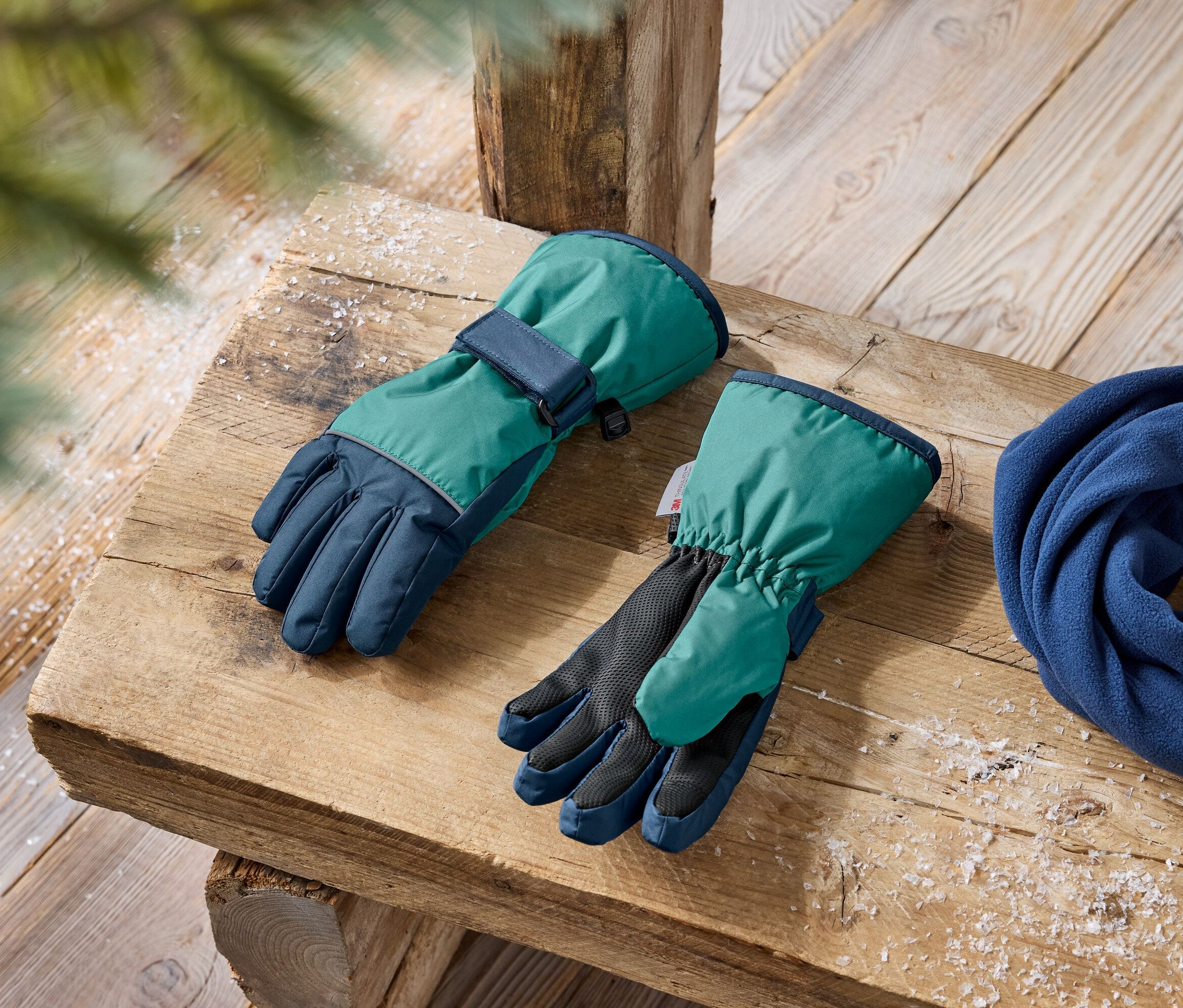 Zimné rukavice v zelenej a tmavomodrej farbe a modrý fleecový šál ležia na drevenej lavici.