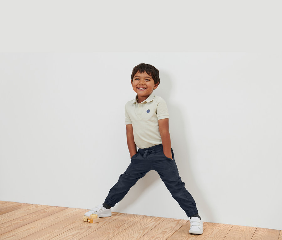 Usmievajúci sa chlapec v béžovej polokošeli, tmavomodrých nohaviciach a bielych topánkach stojí opretý o bielu stenu.