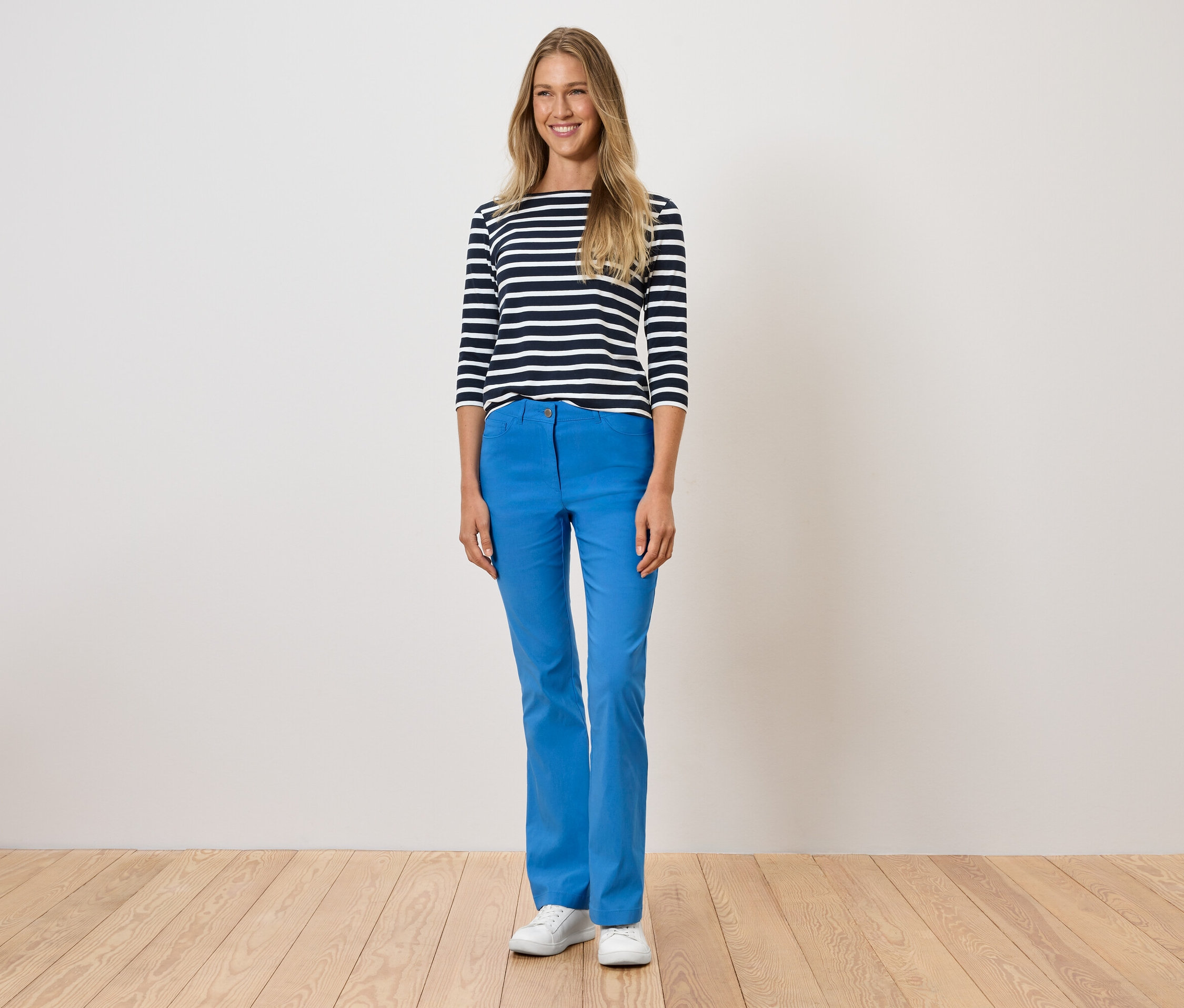Žena stojí a má na sebe modré nohavice, pruhované tričko a biele topánky.