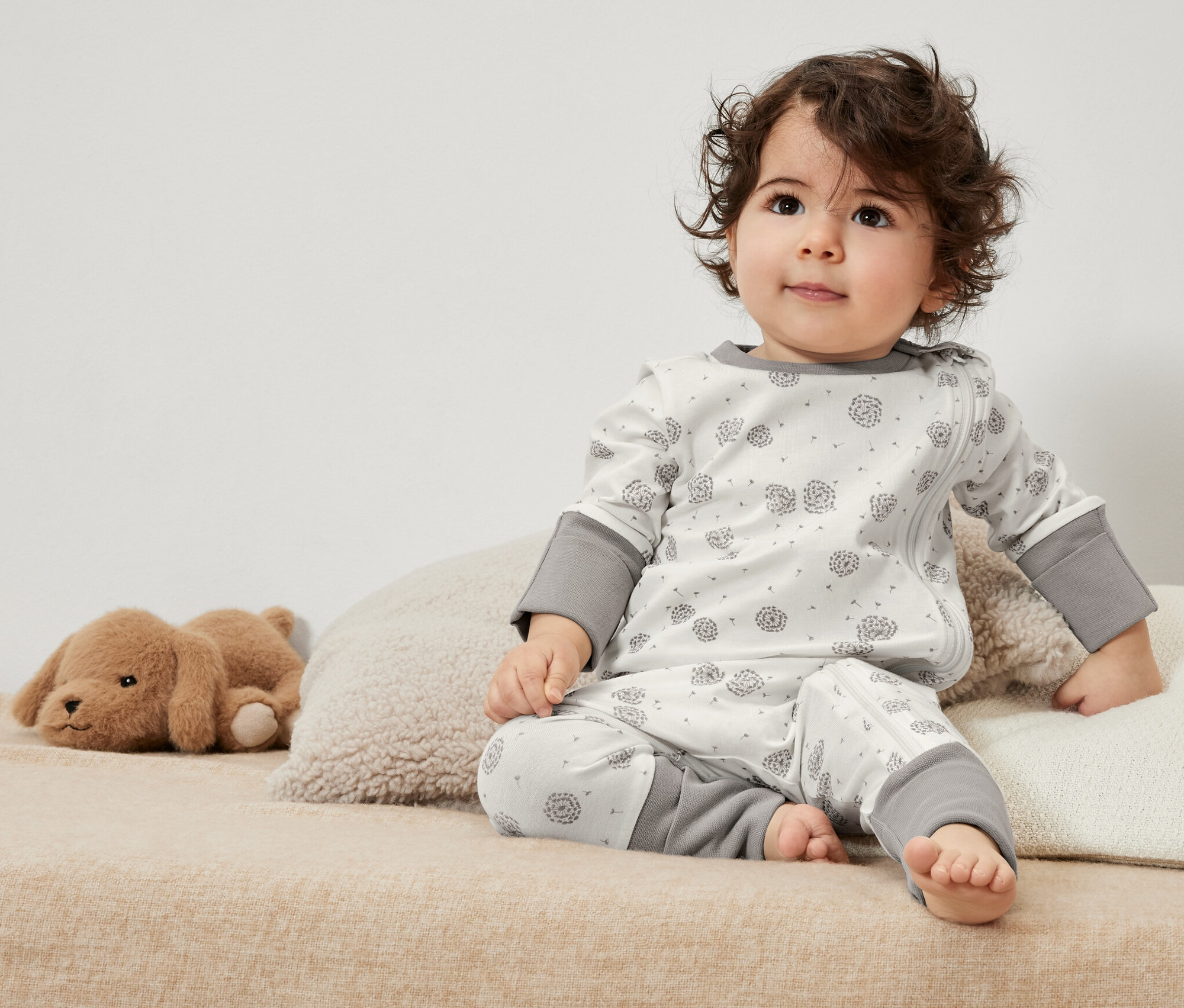 Kučeravé batoľa sedí na posteli v pyžame pre bábätká (3 ks) s hračkou.