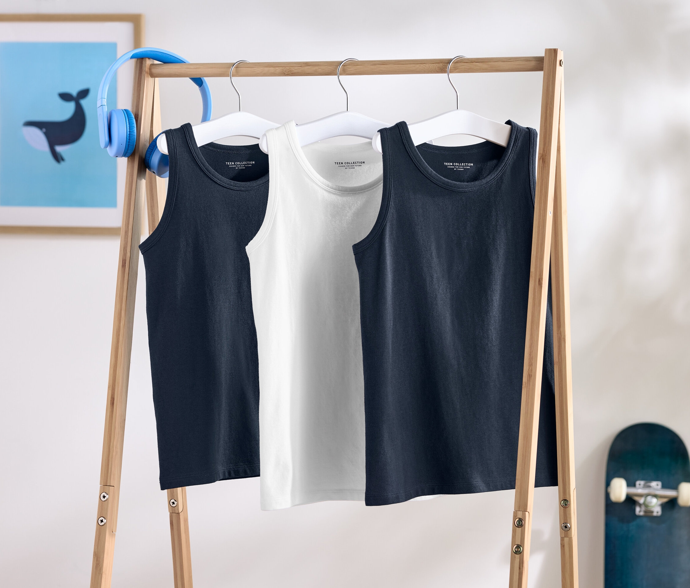 Tričká bez rukávov visia na drevenom stojane, za nimi je skateboard a slúchadlá.