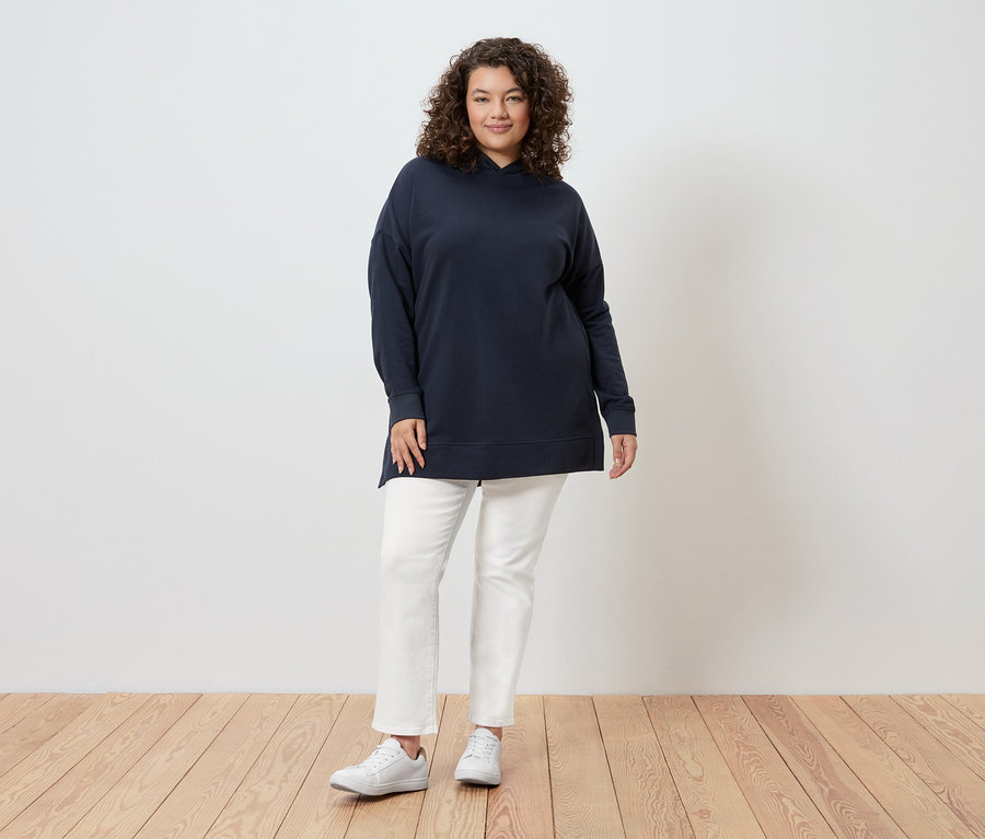 Žena s kučeravými vlasmi stojí pred stenou a má na sebe tmavomodrú mikinu s kapucňou, biele džínsy a biele tenisky.