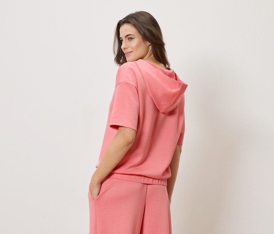 Žena má na sebe koralovú mikinu s krátkym rukávom a zodpovedajúce koralové šortky.