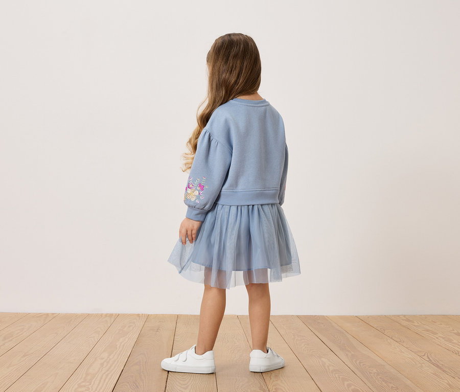 Pohľad zozadu na malé dievča s dlhými hnedými vlasmi, oblečené v modrej mikine s kvetinovým vzorom na rukáve a ladiacej tylovej sukni a bielych teniskách.