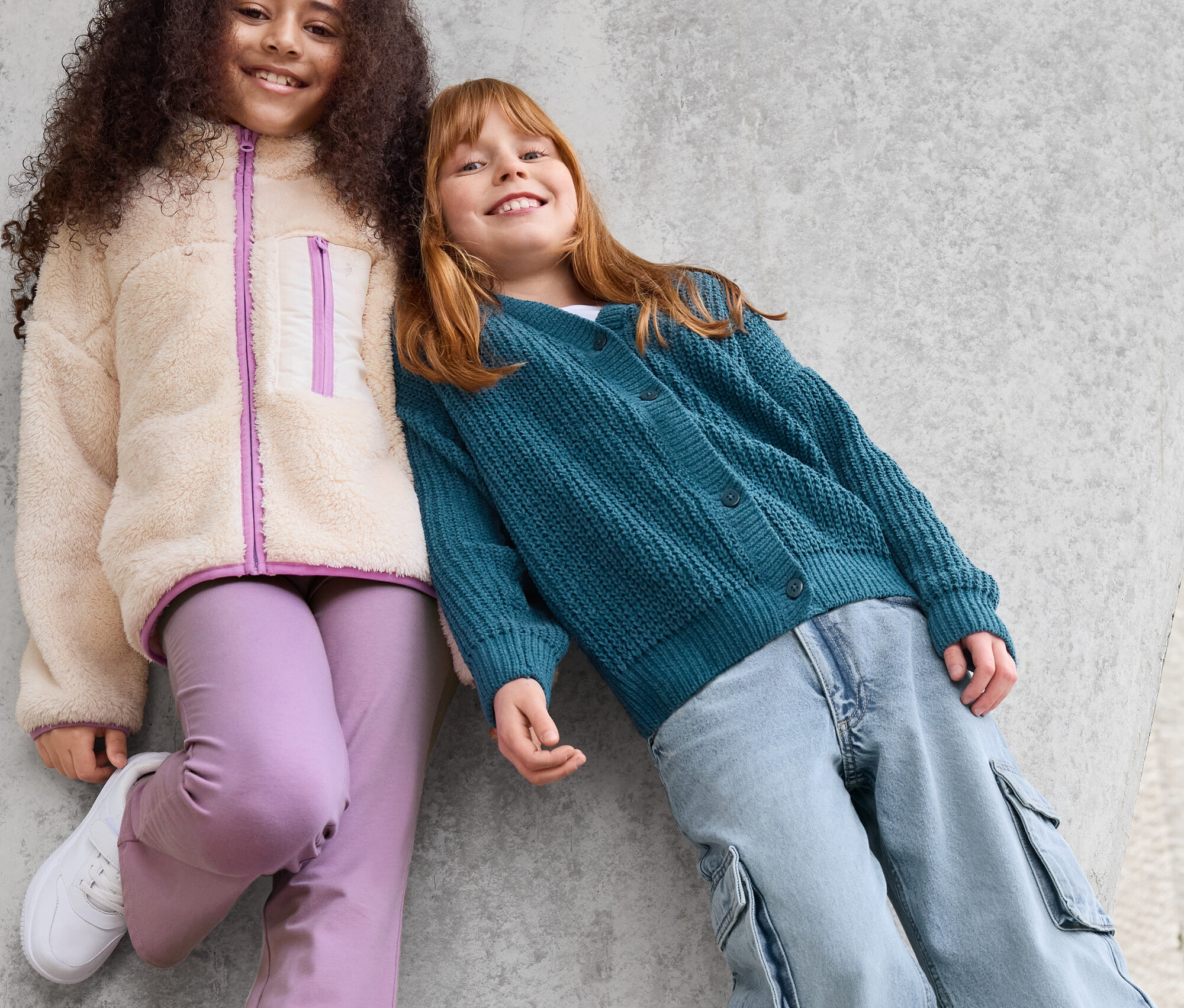 Dve dievčatá sa opierajú o betónovú stenu. Dievča vľavo má na sebe bundu z umelej kožušiny, fialové nohavice a biele tenisky. Dievča vpravo má na sebe zelený kardigán a modré nohavice.