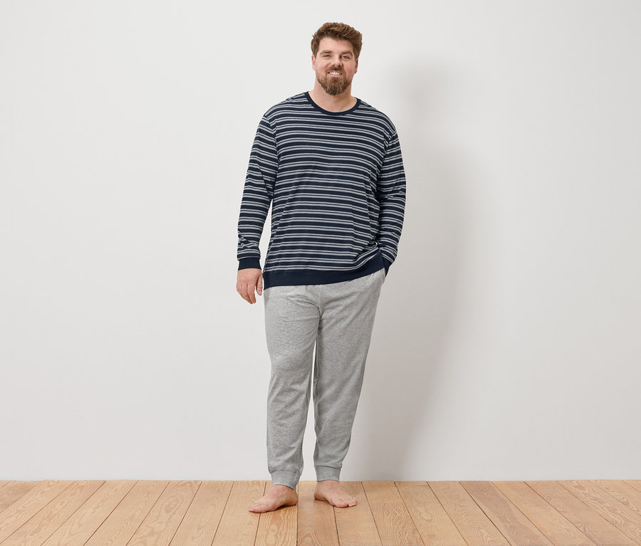 Muž stojí bosý pred stenou a má na sebe sivé pyžamo.