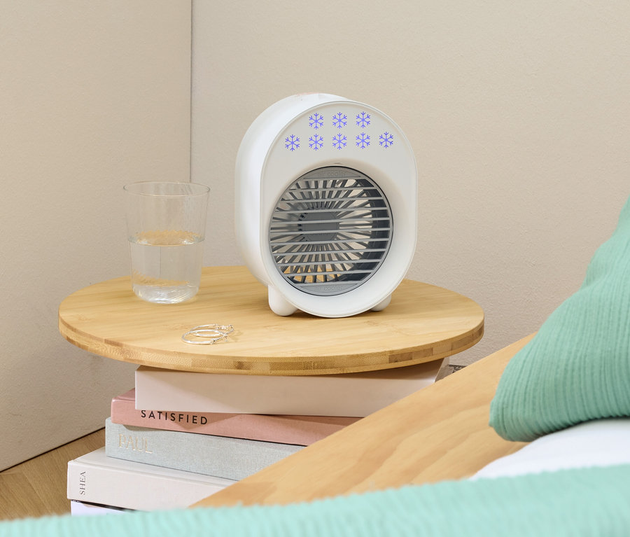 Biely prenosný ventilátor s modrými snehovými vločkami stojí na malom drevenom stolíku vedľa pohára vody a prsteňov.