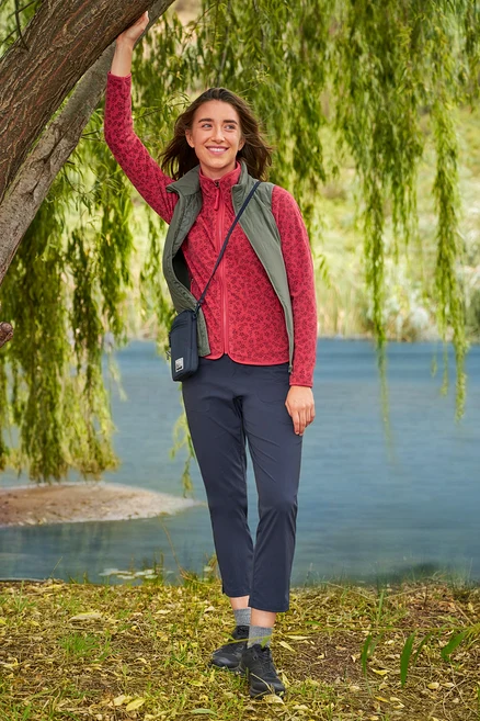 Žena sa opiera o strom pri jazere, má na sebe červenú flísovú bundu, tmavomodré nohavice a khaki vestu »packable« Hybrid-Weste.