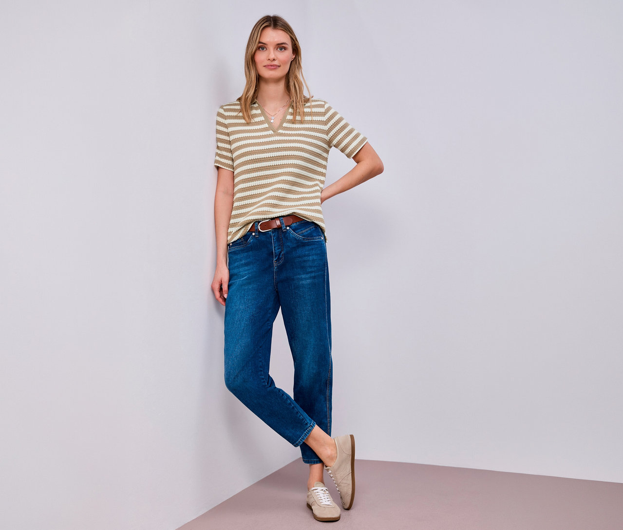 Žena pózuje v tričku z úpletu Street One, džínsoch, hnedom opasku a béžových topánkach v rohu.
