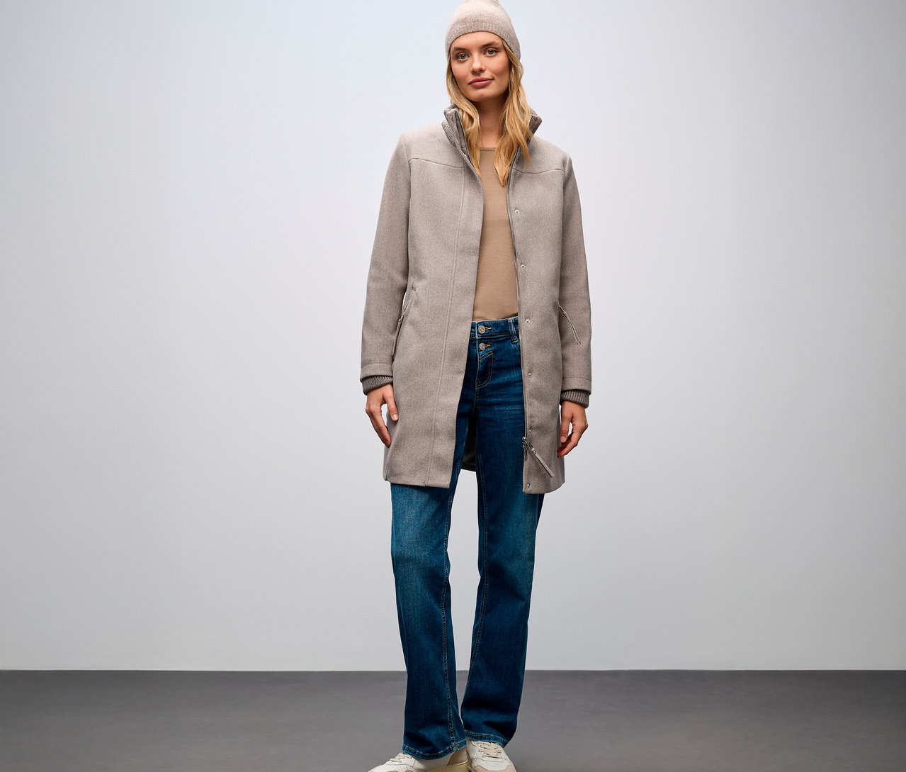 Žena stojí a má na sebe krátky kabát Street One, džínsy a čiapku.