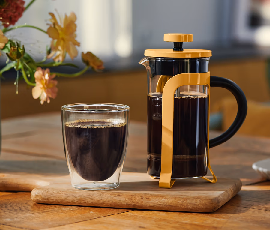 Dvojstenný pohár čiernej kávy a žltý French Press plný kávy stoja na drevenej doske. V pozadí je váza s kvetmi a tanierik s keksíkom.