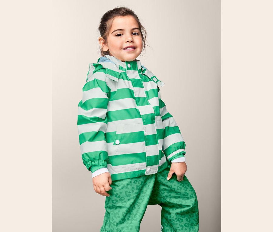 Dievča pózuje v bunde do dažďa z recyklovaného materiálu so zelenými a bielymi pruhmi a zelených nohaviciach s potlačou.