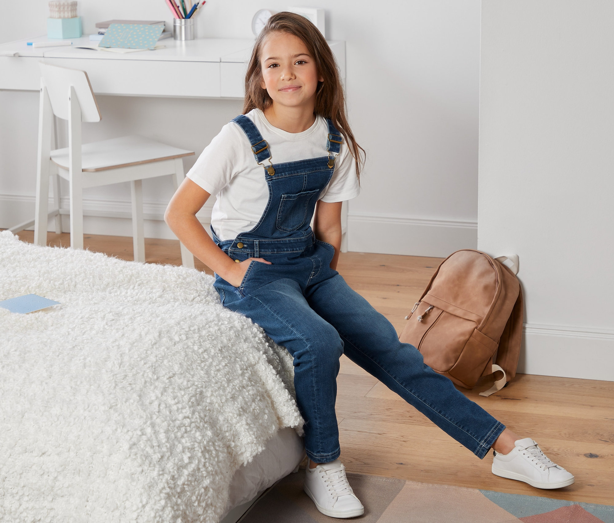 Dievča sedí na posteli a má na sebe džínsy s náprsenkou.