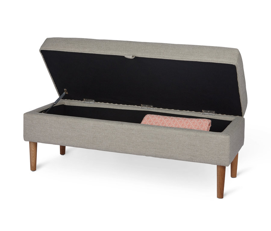 Otvorená čalúnená lavica s úložným priestorom a béžovým poťahom stojí na štyroch drevených nohách. Vnútri je deka s geometrickým vzorom.