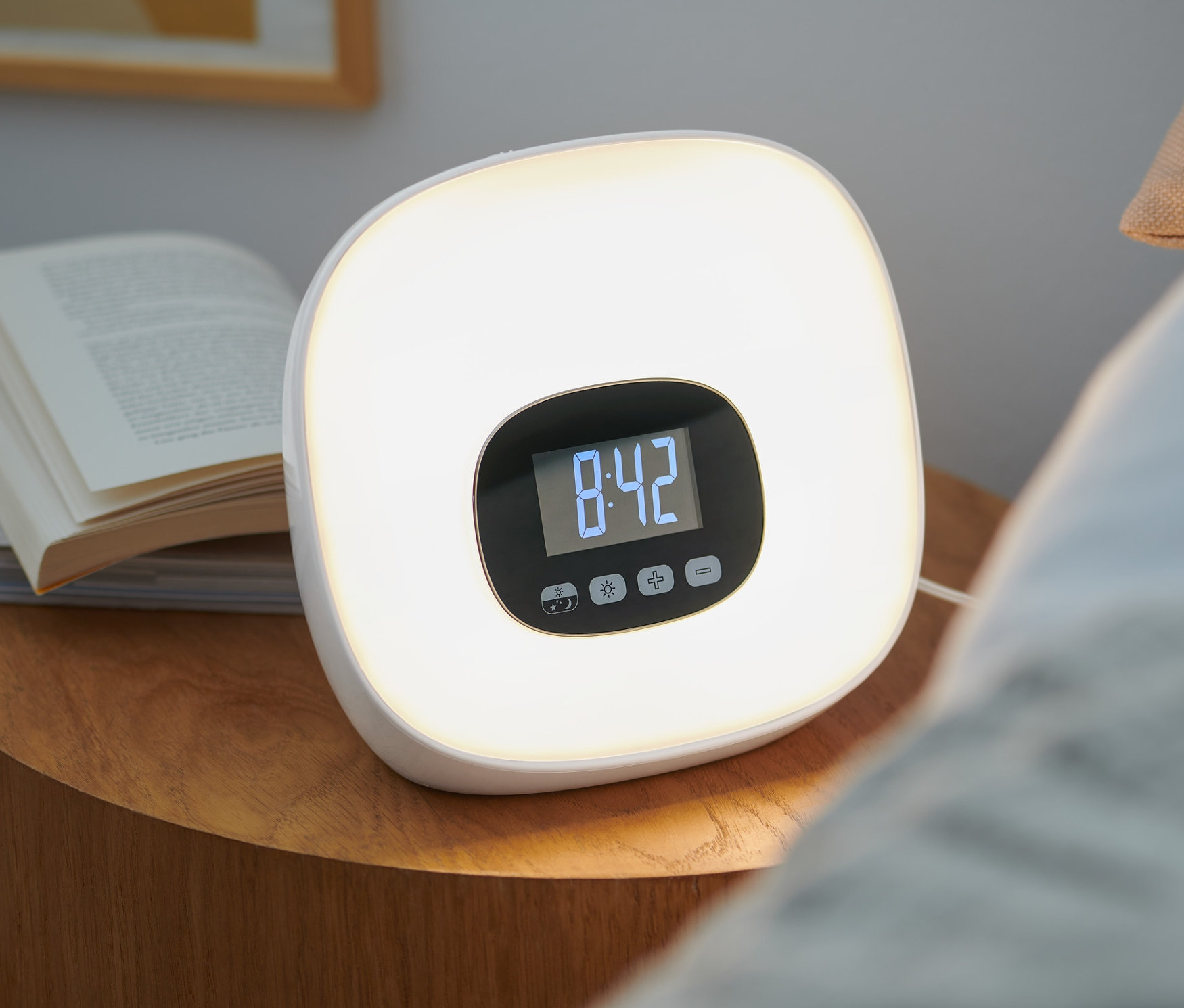 Svetelný budík s rádiom stojí vedľa kníh na nočnom stolíku. Na displeji budíka svieti čas 8:42.