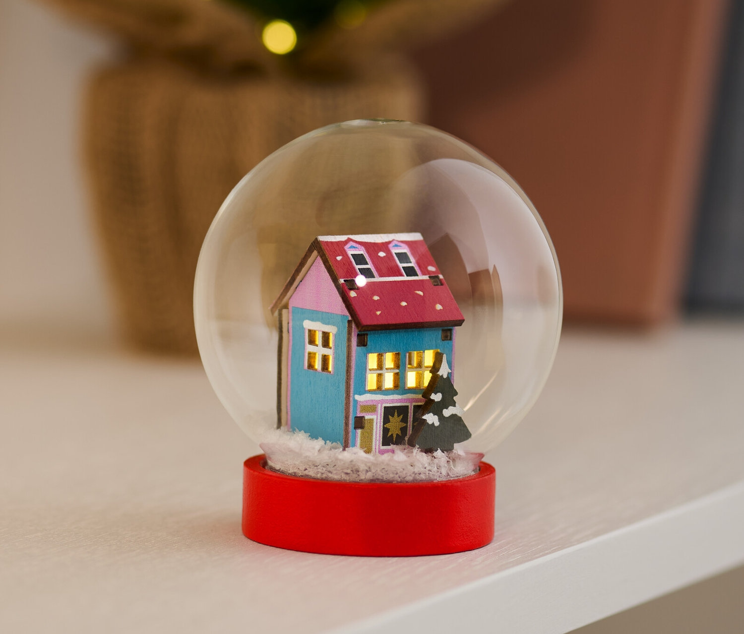 Snehová guľa s LED s modrým domom a červenou základňou stojí na polici.