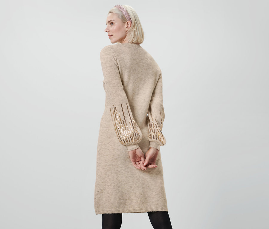 Žena pózuje v béžových úpletových šatách s flitrami a čiernych pančuchách.