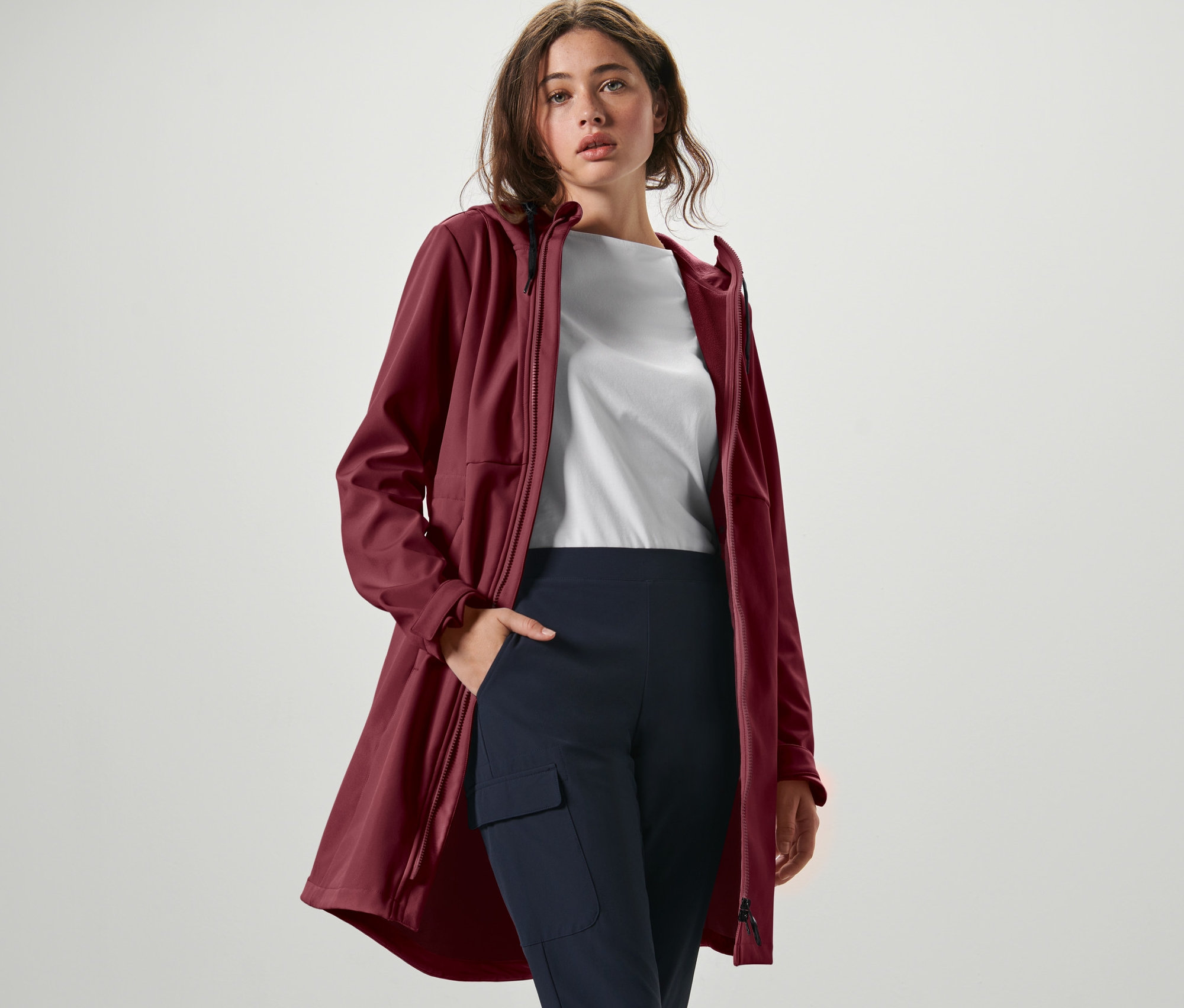 Žena pózuje v softshellovom kabáte vínovo červenej farby, bielom tričku a tmavomodrých nohaviciach.