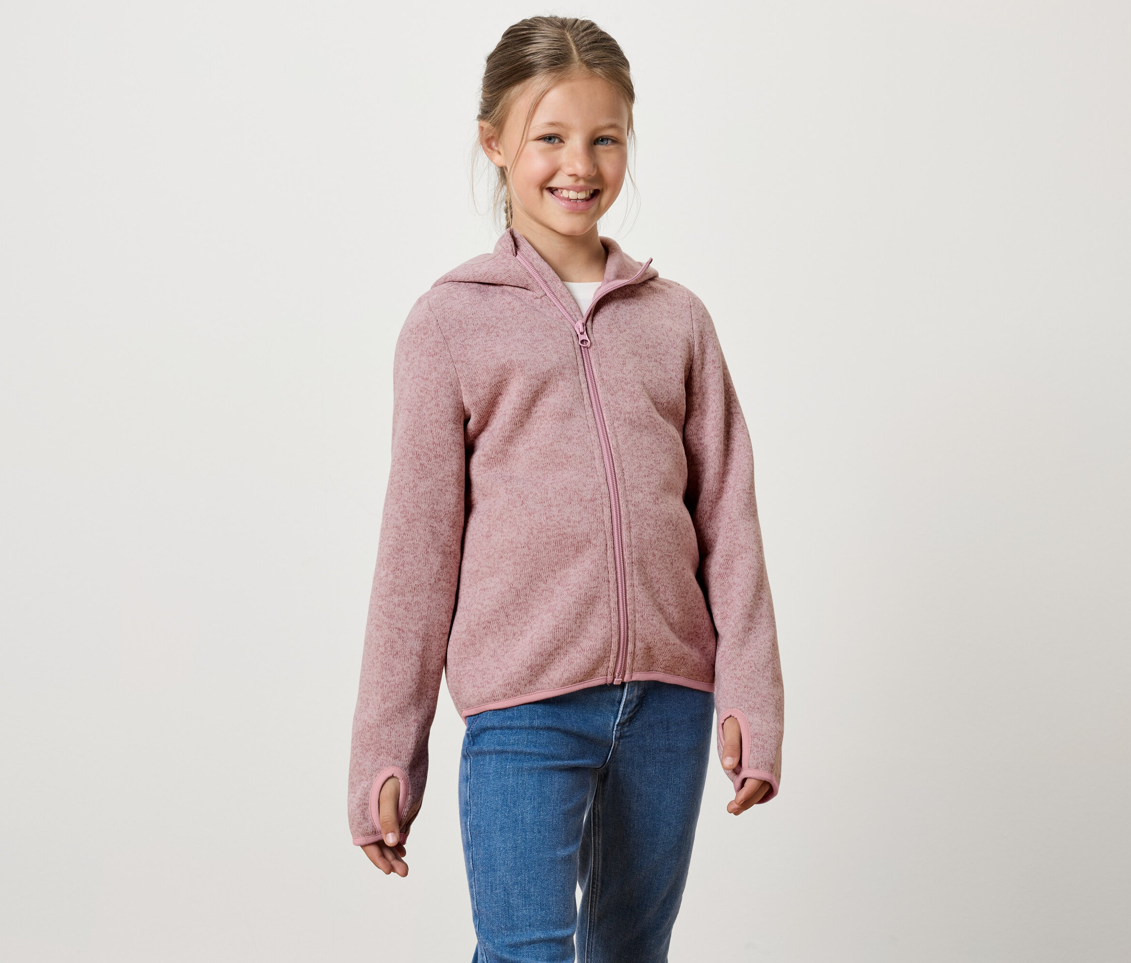 Usmievavé dievča má na sebe ružovú mikinu na zips s kapucňou a modré džínsy.