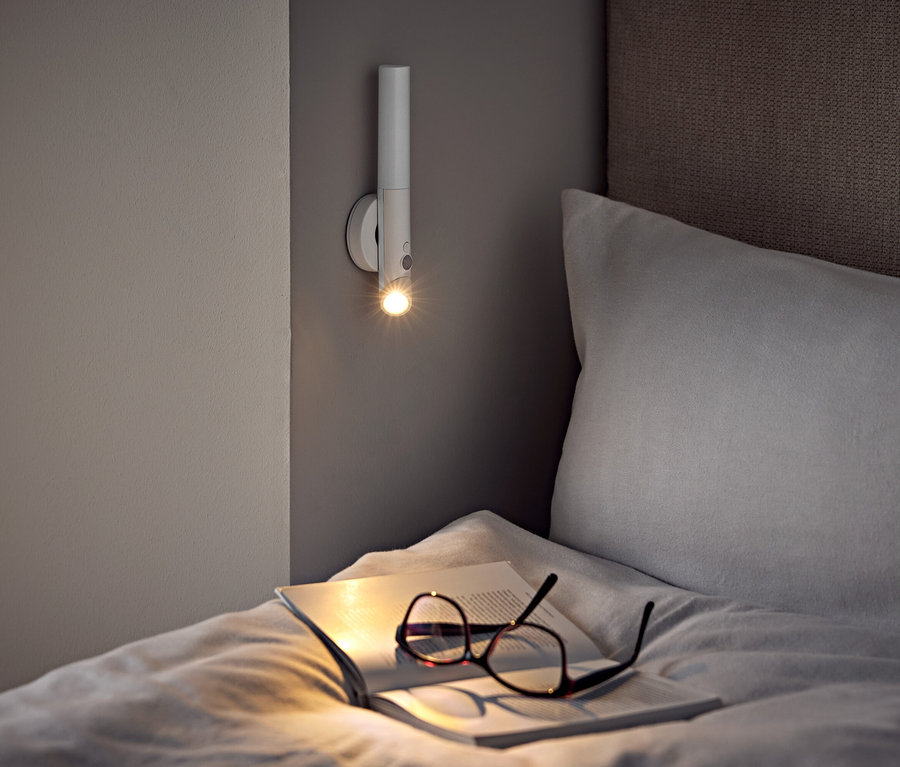Na posteli ležia okuliare a otvorená kniha pod nástennou lampou.