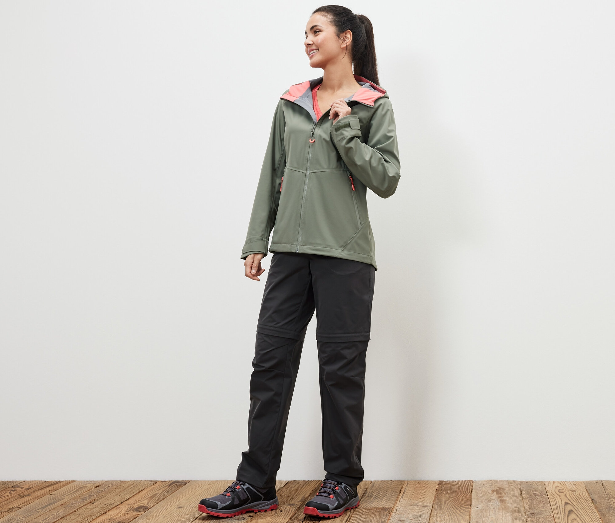 Žena stojaca na drevenej podlahe má na sebe zelenú bundu a čierne funkčné nohavice 2 v 1.