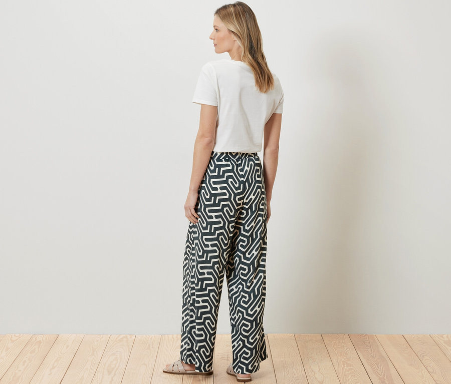 Žena stojí chrbtom k fotoaparátu a má na sebe biele tričko a potlačené pohodlné nohavice v pokrčenom vzhľade.