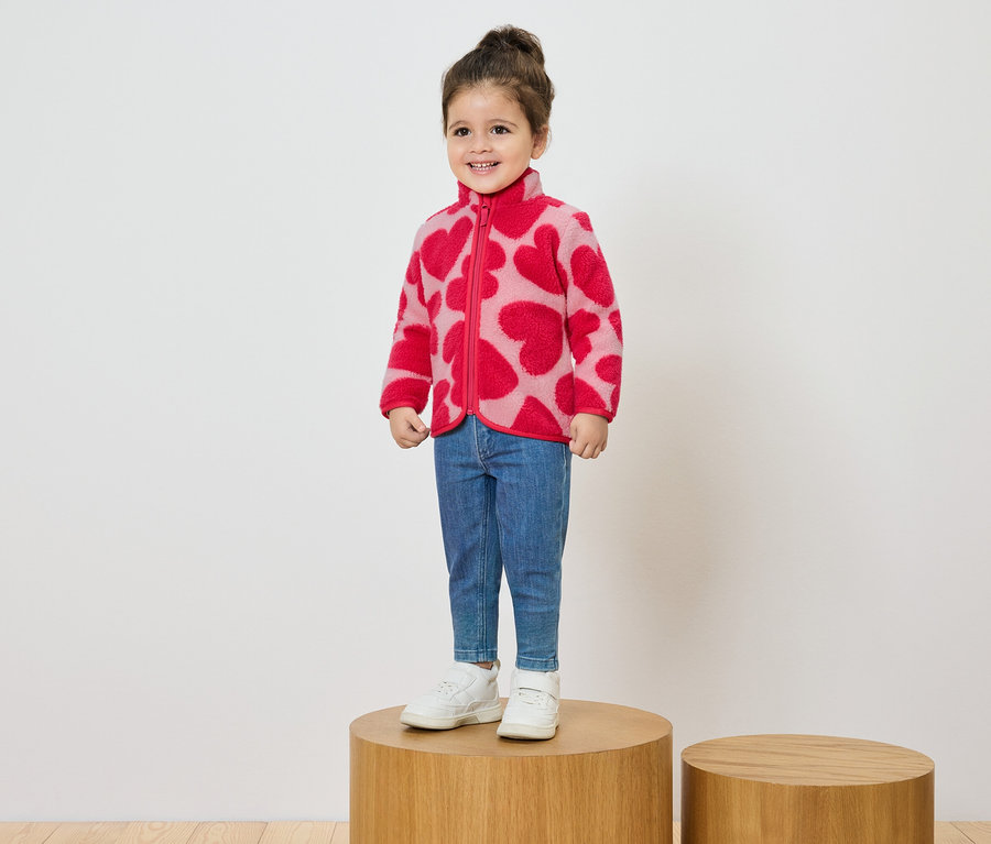 Usmievajúce sa dievčatko stojí na drevenom podstavci a má na sebe ružovú fleecovú bundu so vzorom červených sŕdc, džínsy a biele tenisky.