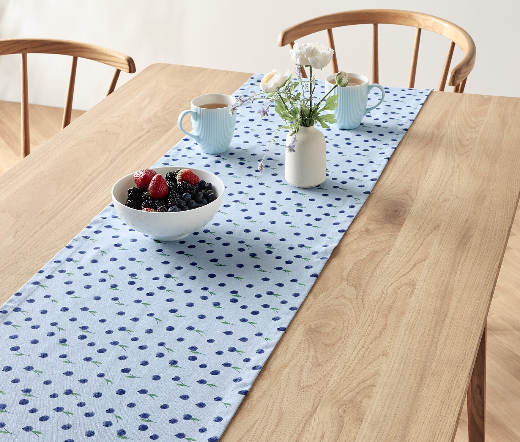 Na stole leží behúň na stôl s čučoriedkovým vzorom, na ňom misa s ovocím, dve modré šálky a váza s kvetmi.
