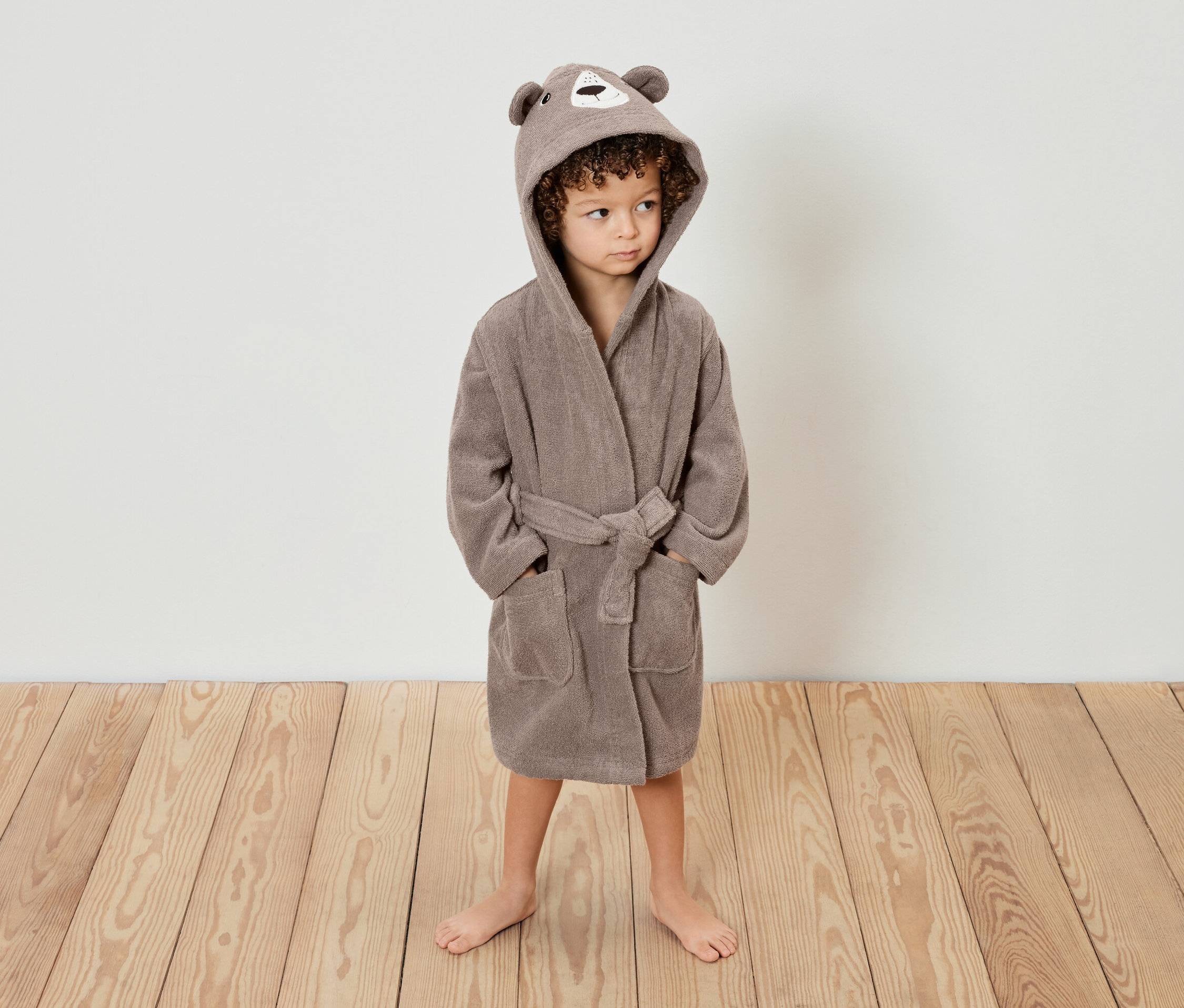 Dieťa s kučeravými vlasmi stojí na drevenej podlahe a má na sebe hnedý župan s kapucňou s uškami medvedíka.