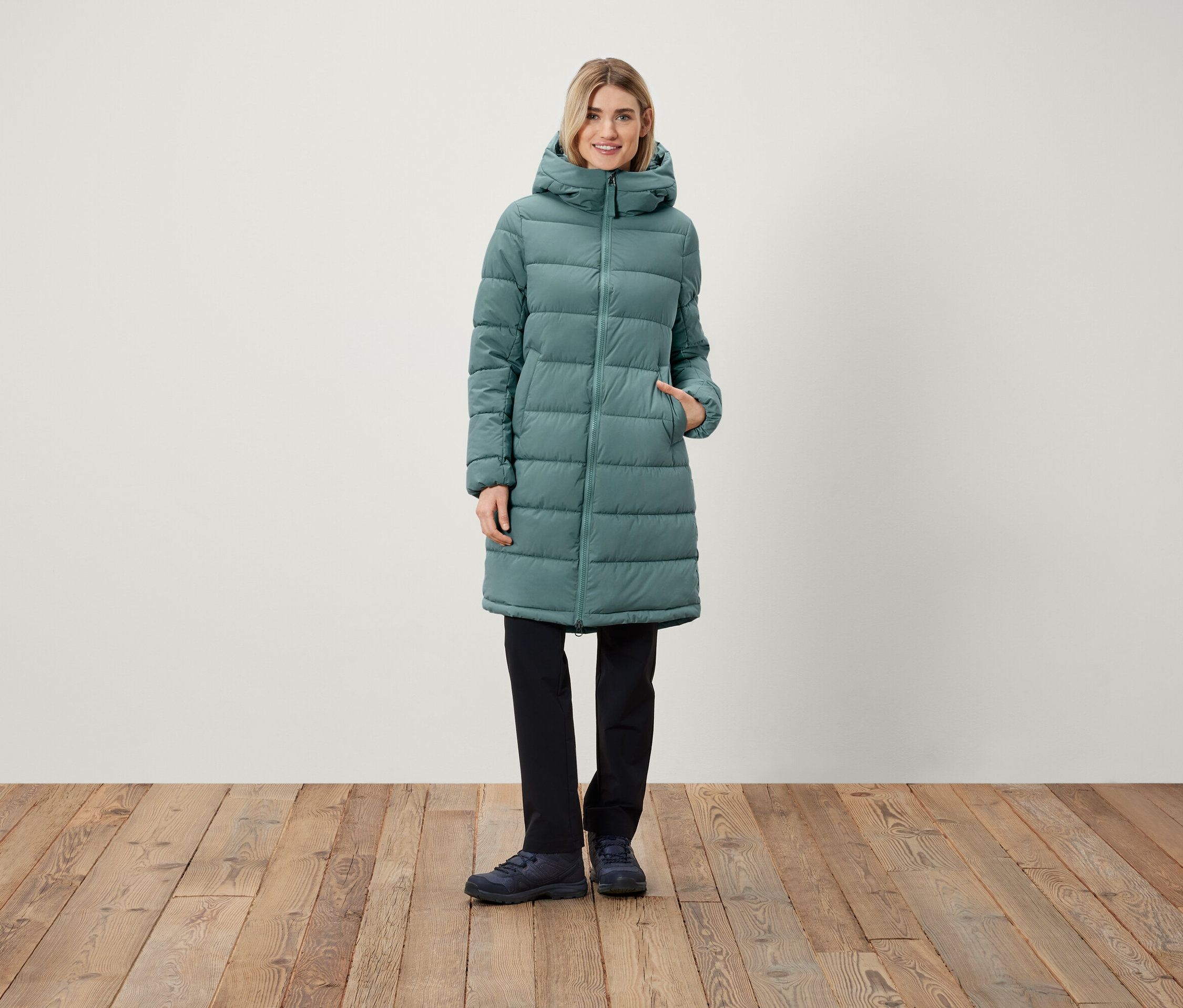 Žena stojí a má na sebe dlhý prešívaný kabát s kapucňou, čierne nohavice a topánky.