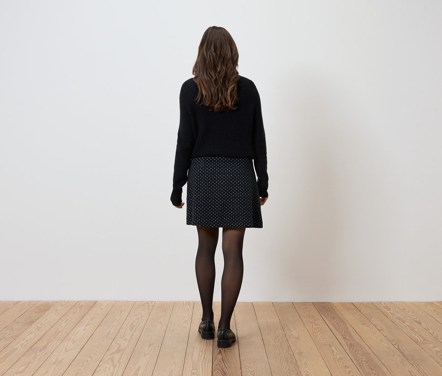 Žena stojí chrbtom k fotoaparátu a má na sebe čierny sveter, vzorovanú sukňu a čierne pančuchy.