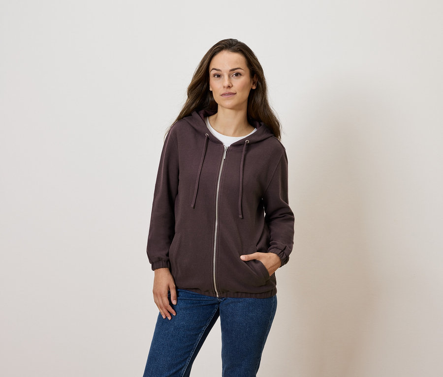 Žena pózuje v hnedej mikine na zips a modrých džínsoch.