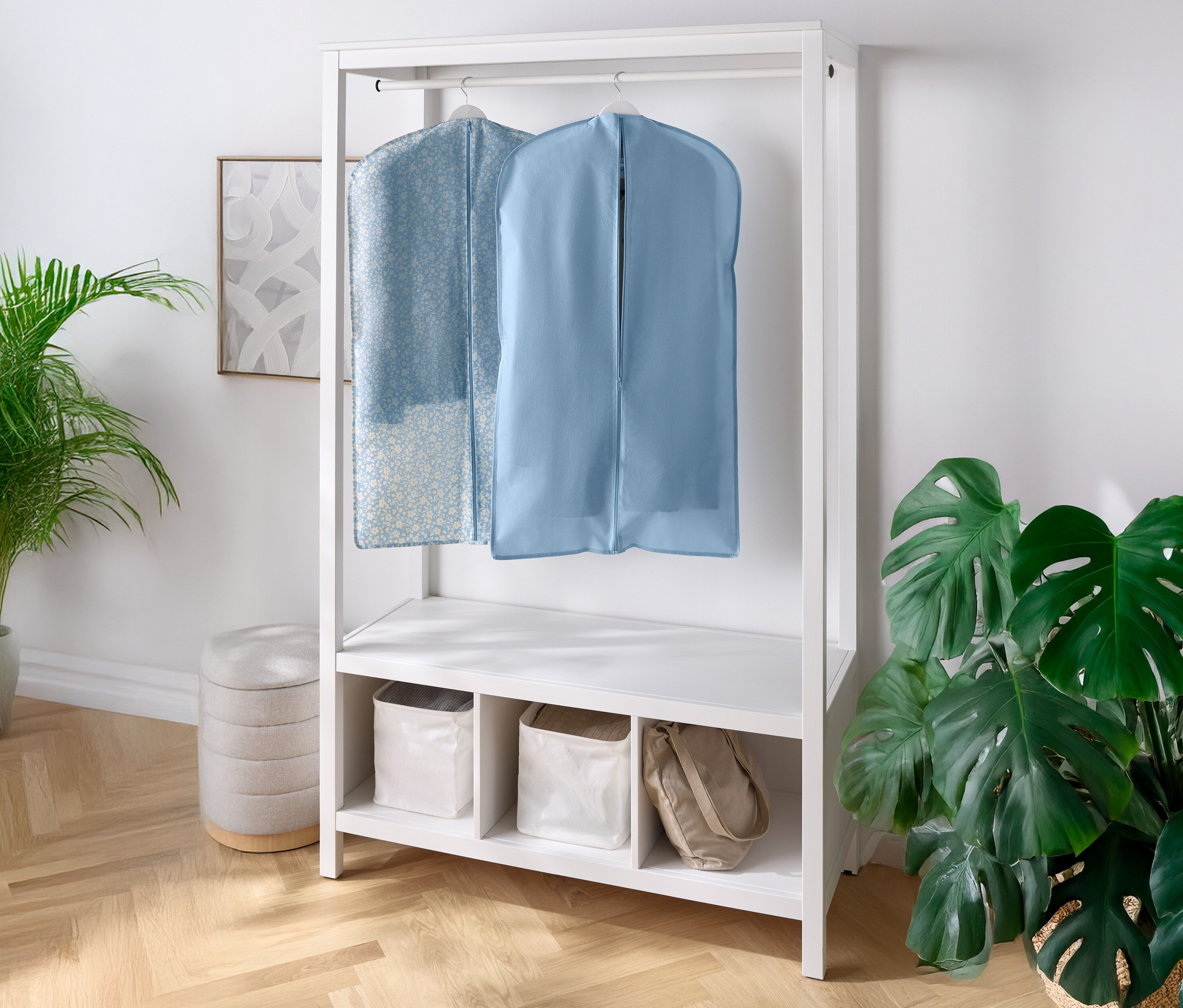 Dva obaly na oblečenie visia na bielom šatníku s policami a priehradkami pod ním. Vľavo a vpravo je izbová rastlina.