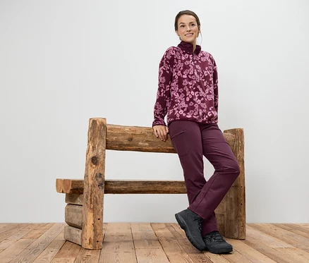 Žena v purpurovom fleecovom svetri s kvetinovou potlačou a nohaviciach sa opiera o drevenú lavičku.
