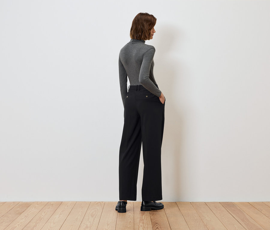 Pohľad zozadu na ženu s rukami vo vreckách čiernych nohavíc a sivým rolákom.