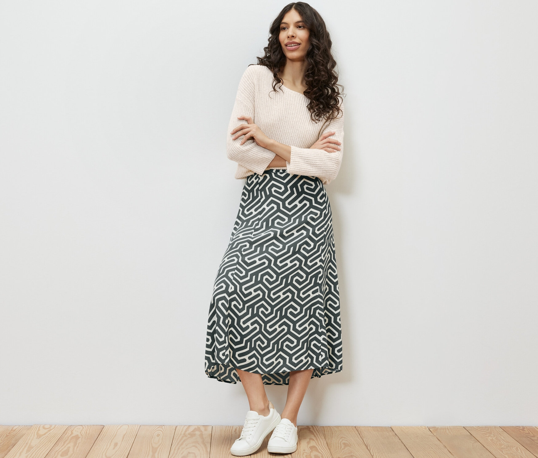 Žena stojí s prekríženými rukami a má na sebe midi sukňu s potlačou, sveter a tenisky.