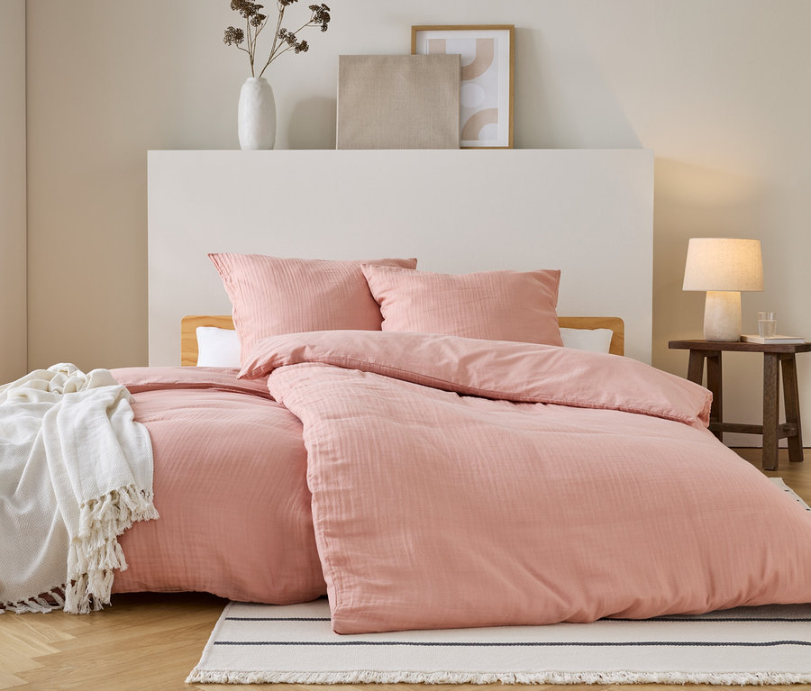Posteľ s ružovou posteľnou bielizňou a vankúšmi stojí na pruhovanom koberci. Na posteli je biela deka. Vedľa stojí nočný stolík.
