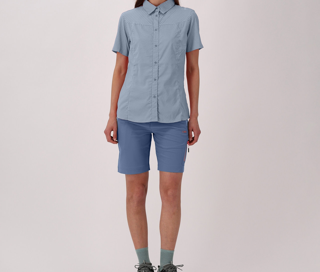 Žena v károvanej outdoorovej blúzke CMP, modrých šortkách, ponožkách a trekových topánkach na svetlom pozadí.