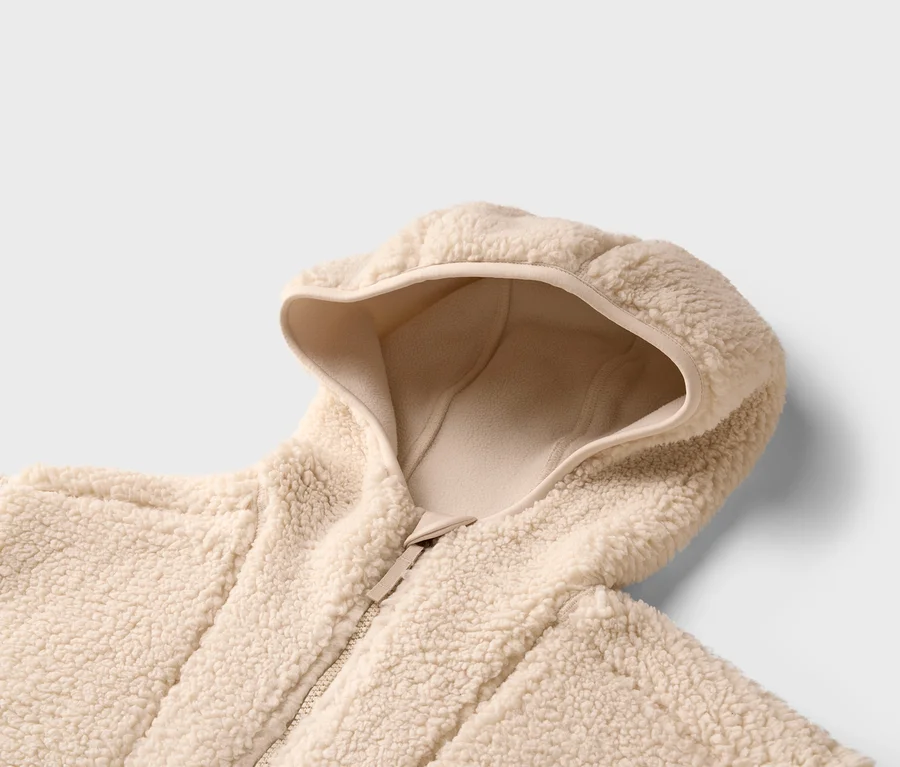 Detail béžovej teddy bundy s kapucňou a zipsom na neutrálnom pozadí.