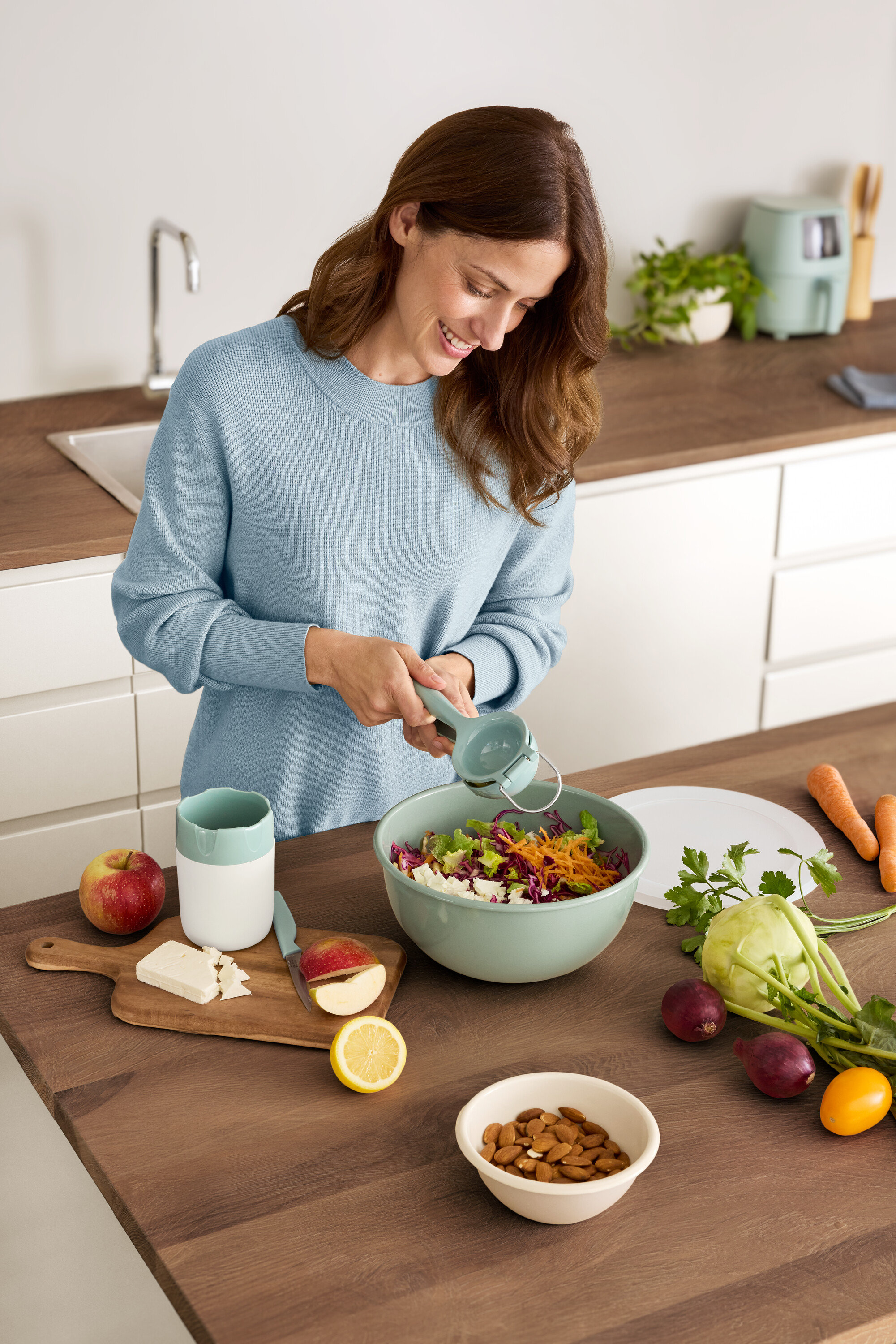 Žena vytláča citrón do šalátovej misy. Na stole ležia jablko, syr, mandle a zelenina.