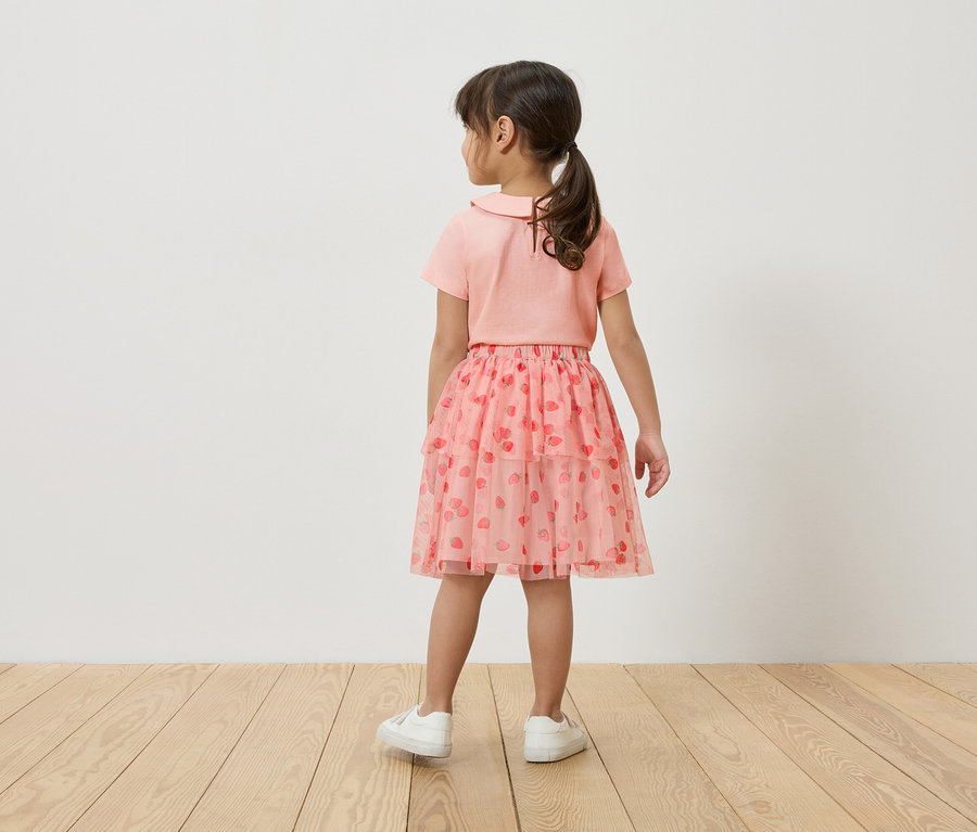 Dievča stojí chrbtom k fotoaparátu, má na sebe ružové tričko, ružovú tylovú sukňu s potlačou jahôd a biele tenisky.